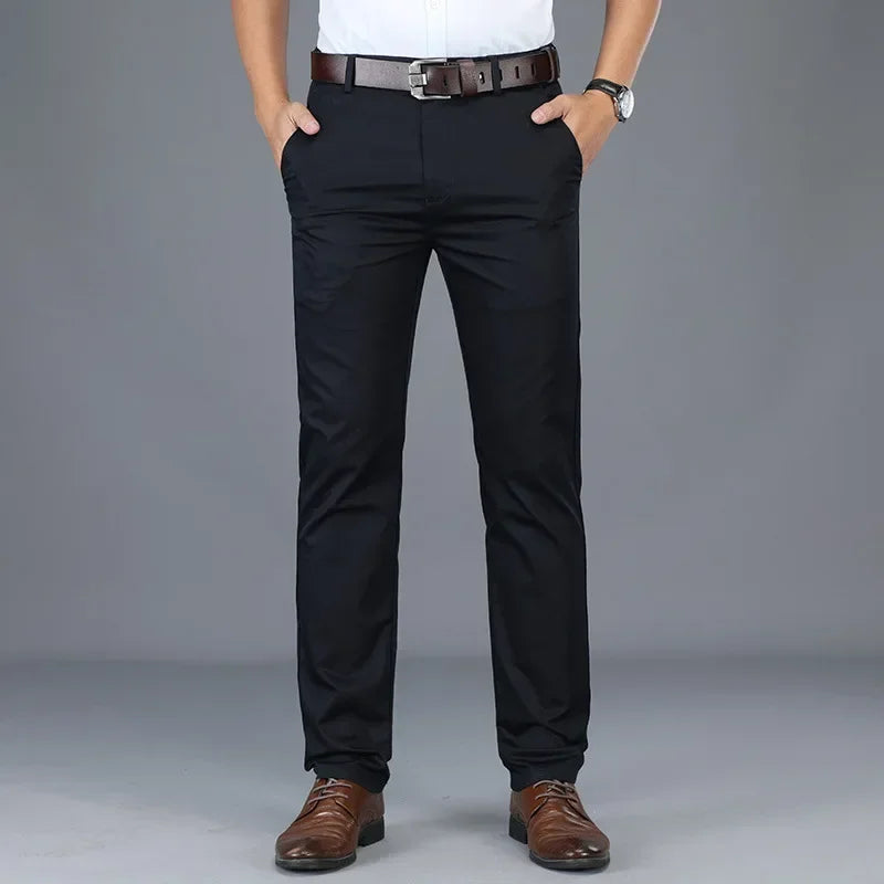 A person wearing a tailored fit white shirt, black belt, black dress pants, and brown dress shoes stands with hands in pockets against a plain gray background. Only the figure from shoulders down is visible.