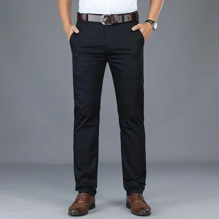A person wearing a tailored fit white shirt, black belt, black dress pants, and brown dress shoes stands with hands in pockets against a plain gray background. Only the figure from shoulders down is visible.