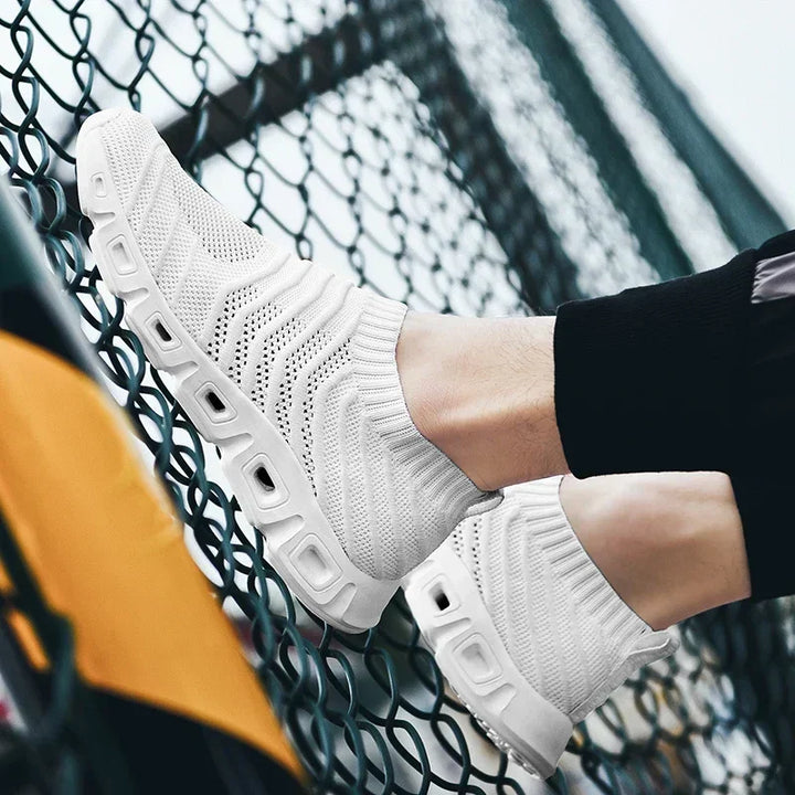 A person wearing white slip-on athletic shoes with a sock-like fit, textured, breathable fabric, and thick, patterned soles rests their feet against a chain-link fence. The person is dressed in black pants.