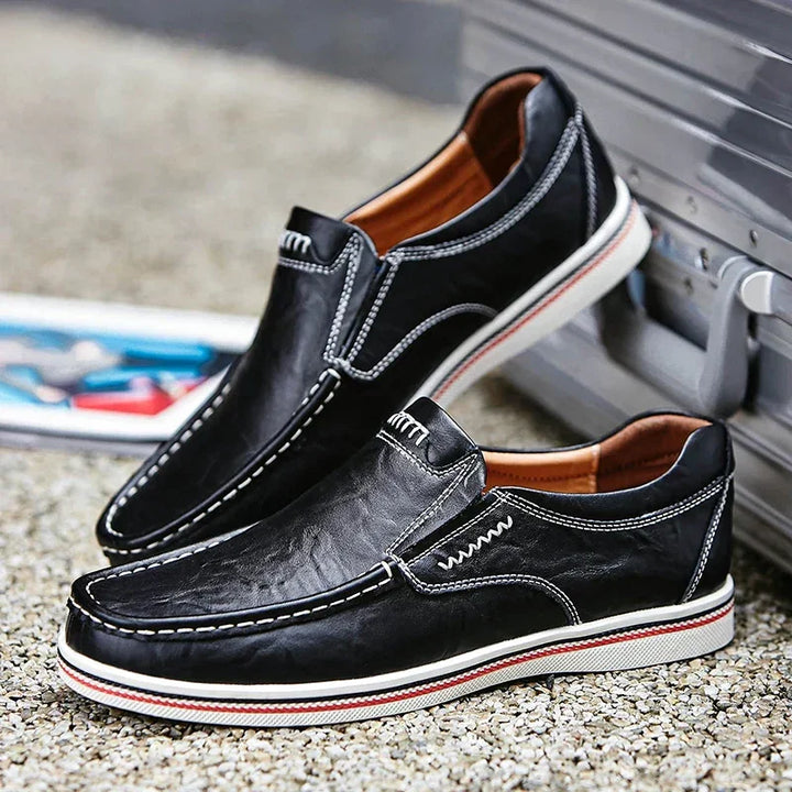 A pair of black leather slip-on shoes with white stitching and white rubber soles, displayed outdoors on a gravel surface next to a silver suitcase.