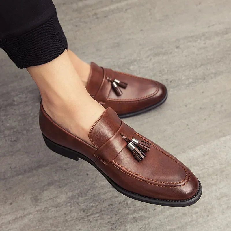 A person wearing polished leather tassel loafers stands on a gray textured floor.