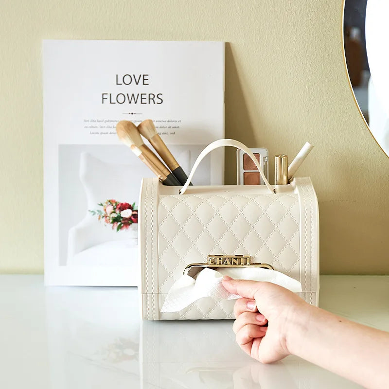 A hand pulls a tissue from a quilted beige fragrance tissue box holder that resembles a handbag. The holder, perfect for home decor, sits on a white desk with makeup brushes and pens, next to a magazine titled Love Flowers.