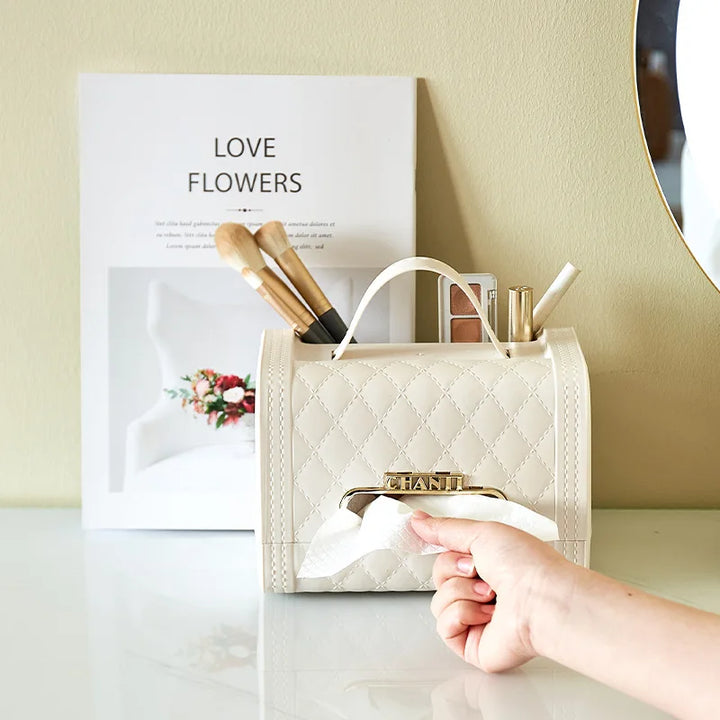 A hand pulls a tissue from a quilted beige fragrance tissue box holder that resembles a handbag. The holder, perfect for home decor, sits on a white desk with makeup brushes and pens, next to a magazine titled Love Flowers.