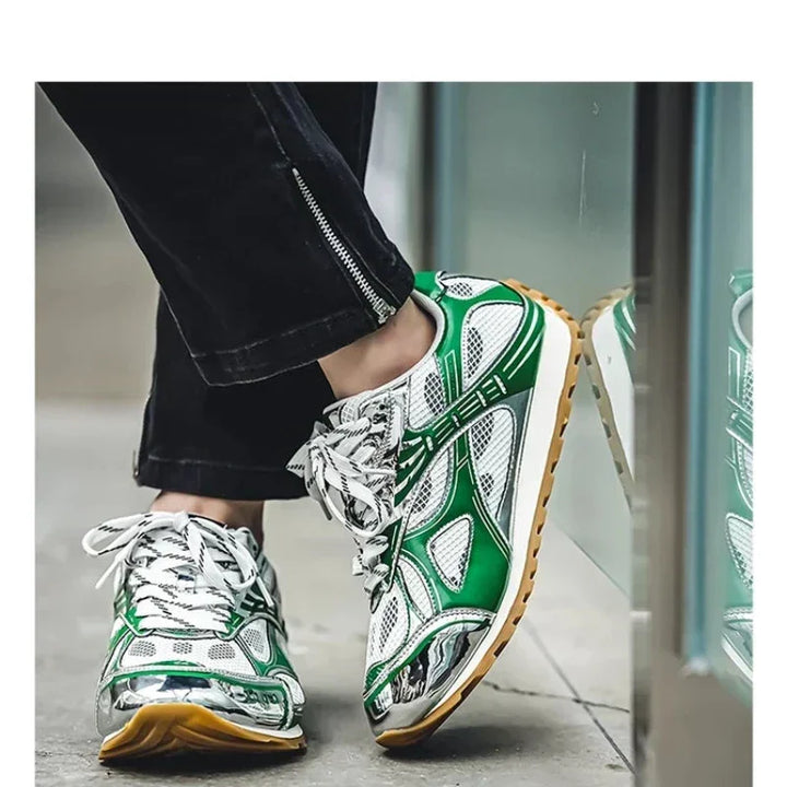 A person wearing black pants and sporty sneakers with green, white, and silver details stands near a glass surface. The lightweight mesh shoes feature patterned laces, a cushioned insole, and tan-colored rubber soles.