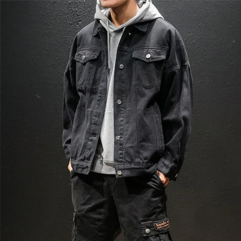 A person wearing a light gray hoodie layered under a black premium denim jacket and black cargo pants stands against a plain dark background. The persons head is partially out of frame.
