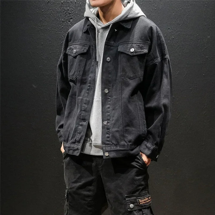 A person wearing a light gray hoodie layered under a black premium denim jacket and black cargo pants stands against a plain dark background. The persons head is partially out of frame.
