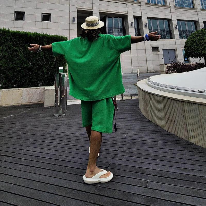 Person in green oversized shirt and shorts, straw hat, and sandals outdoors by modern building