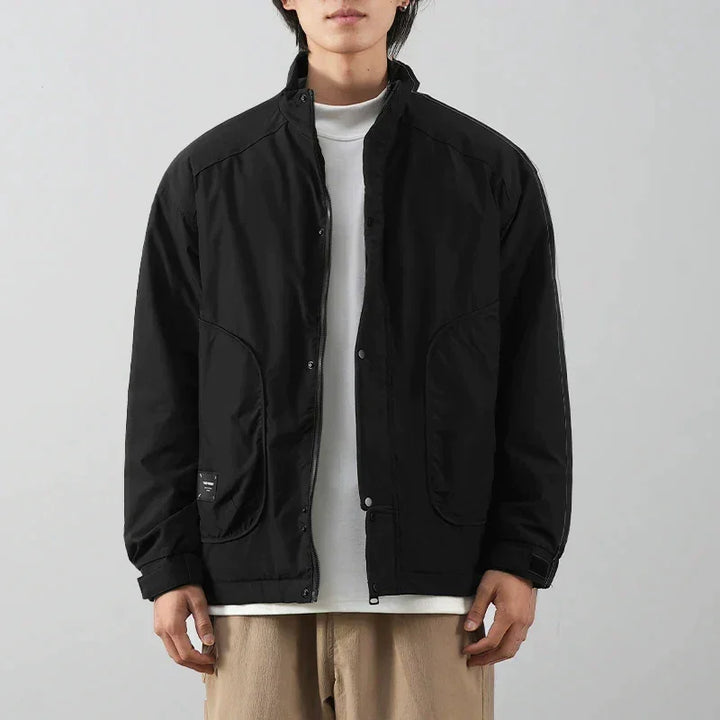 A person wearing a minimalist jacket with a black exterior over a white t-shirt and beige pants stands against a plain light gray background. The individuals face is partially out of frame.