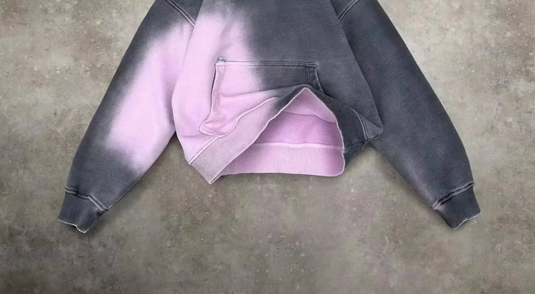 A cropped hoodie with a relaxed fit and front pocket, featuring a two-tone design in dark gray and light pink, laid flat on a textured gray surface.