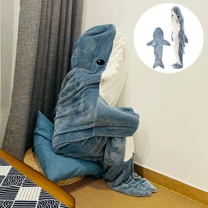 A person sits on a cushion, curled up and wearing cozy shark pajamas with a fin, tail, and face. An inset shows the same plush blue and white costume being worn while standing.