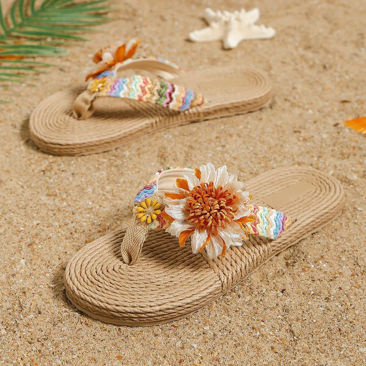 A pair of beige woven sandals with colorful straps and large fabric flowers, designed as a women's fashion accessory, rests on sandy beach with a seashell and a palm leaf nearby.