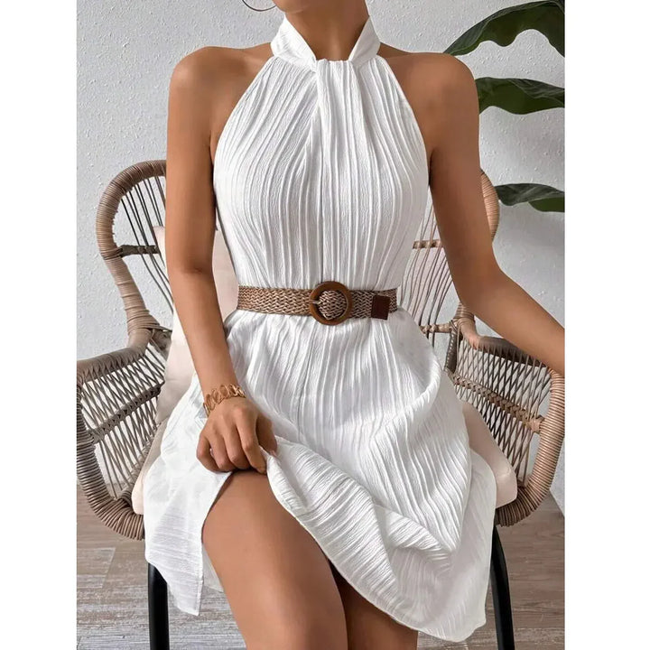 Woman in pleated white halter dress with woven belt, seated on rattan chair, Rovelli fashion