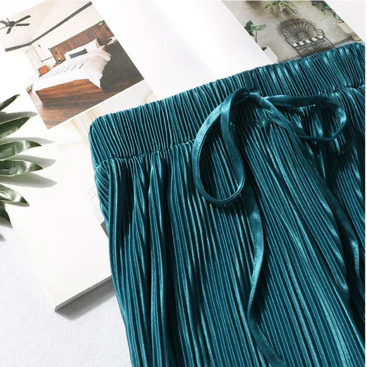 A pair of teal, pleated ice silk women fashion summer pants with a drawstring waistband is laid on an open magazine featuring bedroom and chair images. Green leaves are partially visible beside the wide leg pants.