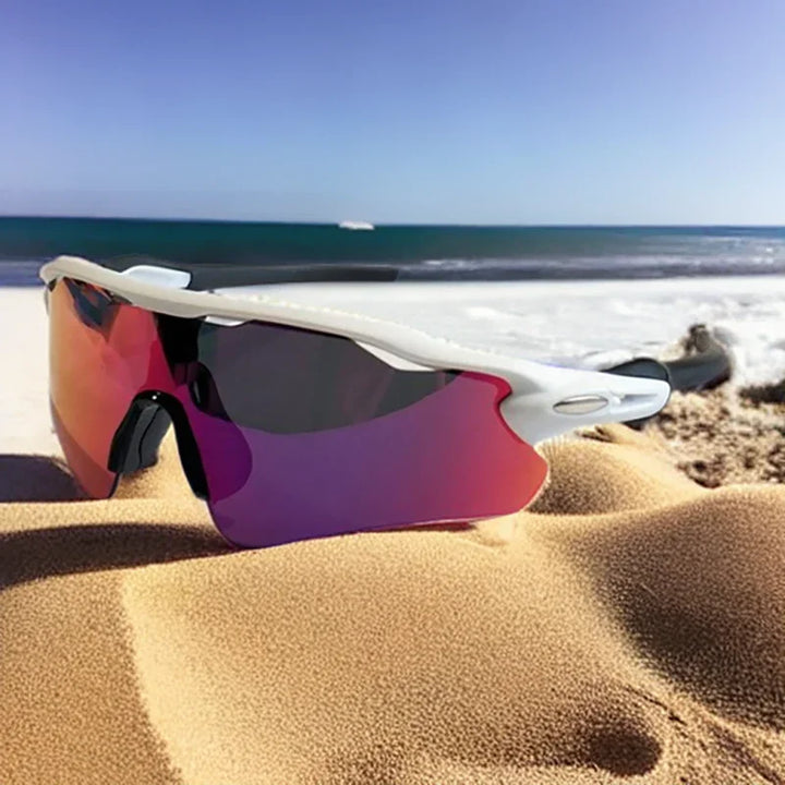 A pair of white-framed, mirrored Polarized Cycling Sunglasses with UV Protection resting on sandy beach with the ocean and blue sky in the background.