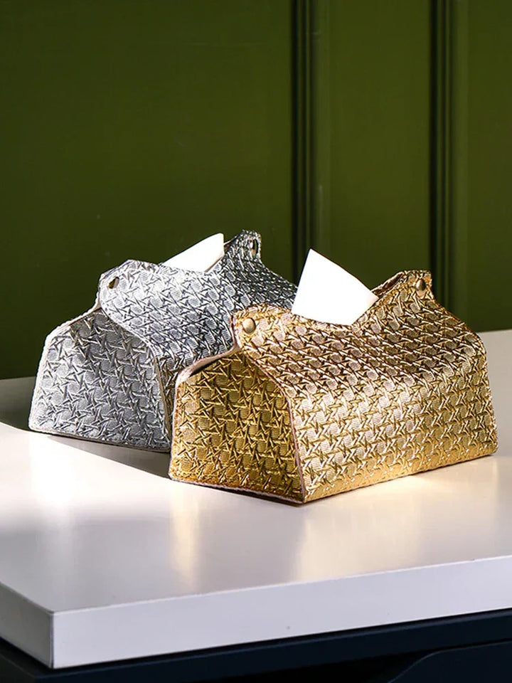 Two decorative tissue holders, including a Glymmer Tissue Box in metallic gold and a silver geometric tissue box, sit side by side on a white table against a green background. A tissue is partially pulled out from the gold holder.