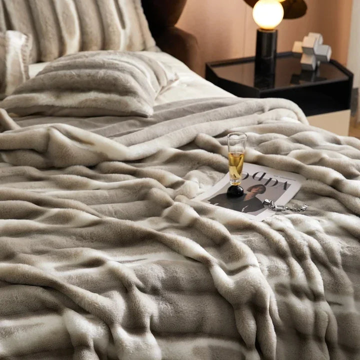 A cozy bed with a soft, striped, gray and white faux rabbit fur blanket, matching pillows, a magazine, eyeglasses, and a glass of drink on top. A modern nightstand with a lamp and decorative objects sits beside the bed.