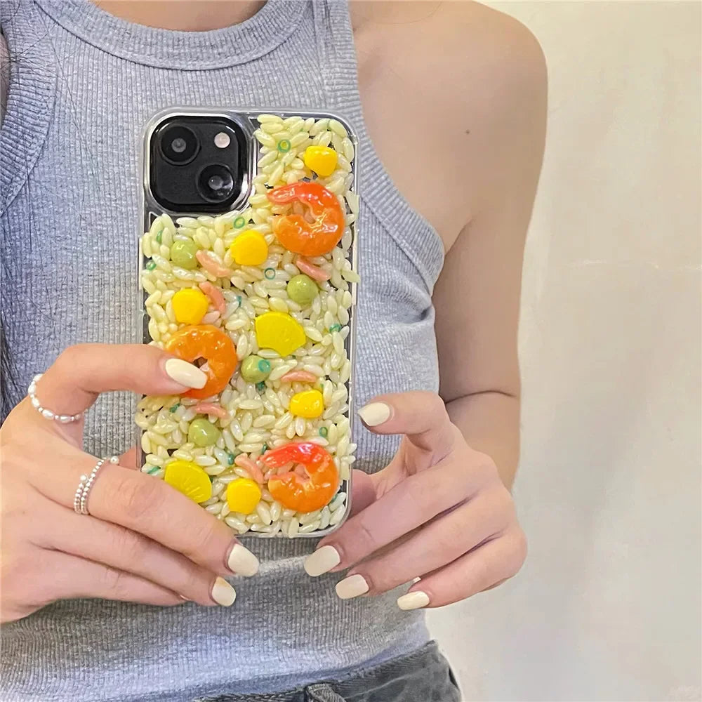 A person holds a phone with a case designed to look like fried rice, featuring realistic 3D detailing of shrimp, corn, peas, and other colorful toppings. The rugged phone protection adds durability while the person shows off manicured nails and a sleeveless gray top.