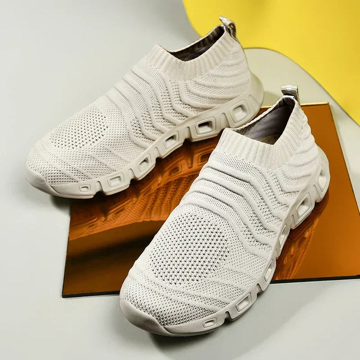 A pair of modern, slip-on knit sneakers in light beige with a sock-like fit and thick, cushioned soles, featuring textured patterns. Displayed on a reflective brown surface with a yellow accent in the background.