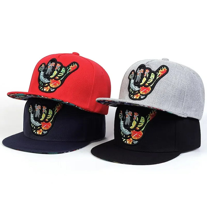Four snapback hats with a colorful embroidered shaka hand symbol on the front. Each Hawaiian-inspired cap comes in red, gray, navy blue, or black, featuring a vibrant floral pattern under the brim for that tropical snapback vibe.