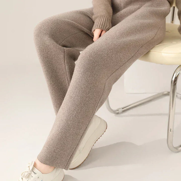 A person sitting on a chair, wearing a beige knit sweater and slim tailored leg pants with white sneakers, shown from the shoulders down.