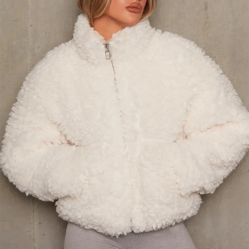 A person wearing a fluffy, white sherpa jacket with a full zip closure and hands in the pockets stands against a plain light gray wall. Their face is partially visible, and they are dressed in light gray pants. The look is warm weightless and cozy.