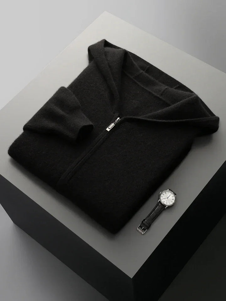 A neatly folded black men wool cardigan and a black leather-strapped wristwatch with a white face are displayed on a square, light gray surface against a neutral background.