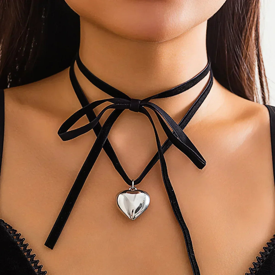 A woman wears a black velvet choker tied in a bow, featuring a vintage heart pendant in shiny silver. She pairs the adjustable choker with a black top trimmed in lace.