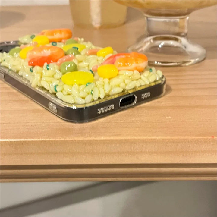 A phone with a shock-absorbent case designed to look like rice and colorful vegetables sits on a wooden surface near a glass dessert dish, featuring playful 3D detailing for added charm.