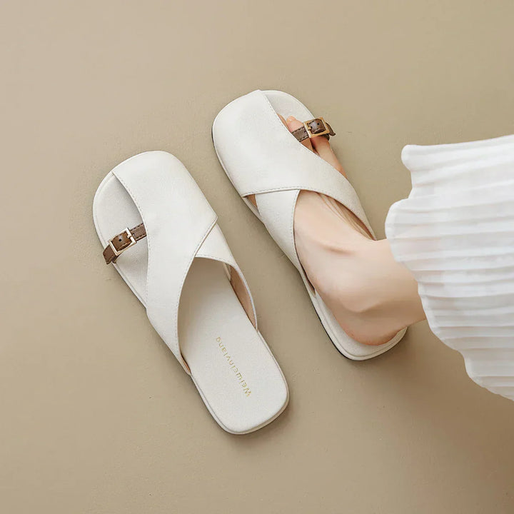A person wears chic leather slides with crisscross straps and small buckles, shown on a beige floor, with part of a white pleated skirt visible—a stylish addition to any summer wardrobe.