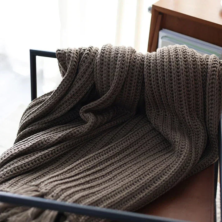 A chunky weave throw in brown is draped over the back of a black metal chair, with a wooden shelf and books visible in the background. Soft daylight filters in from the left side, highlighting this cozy, stress reducing blanket.
