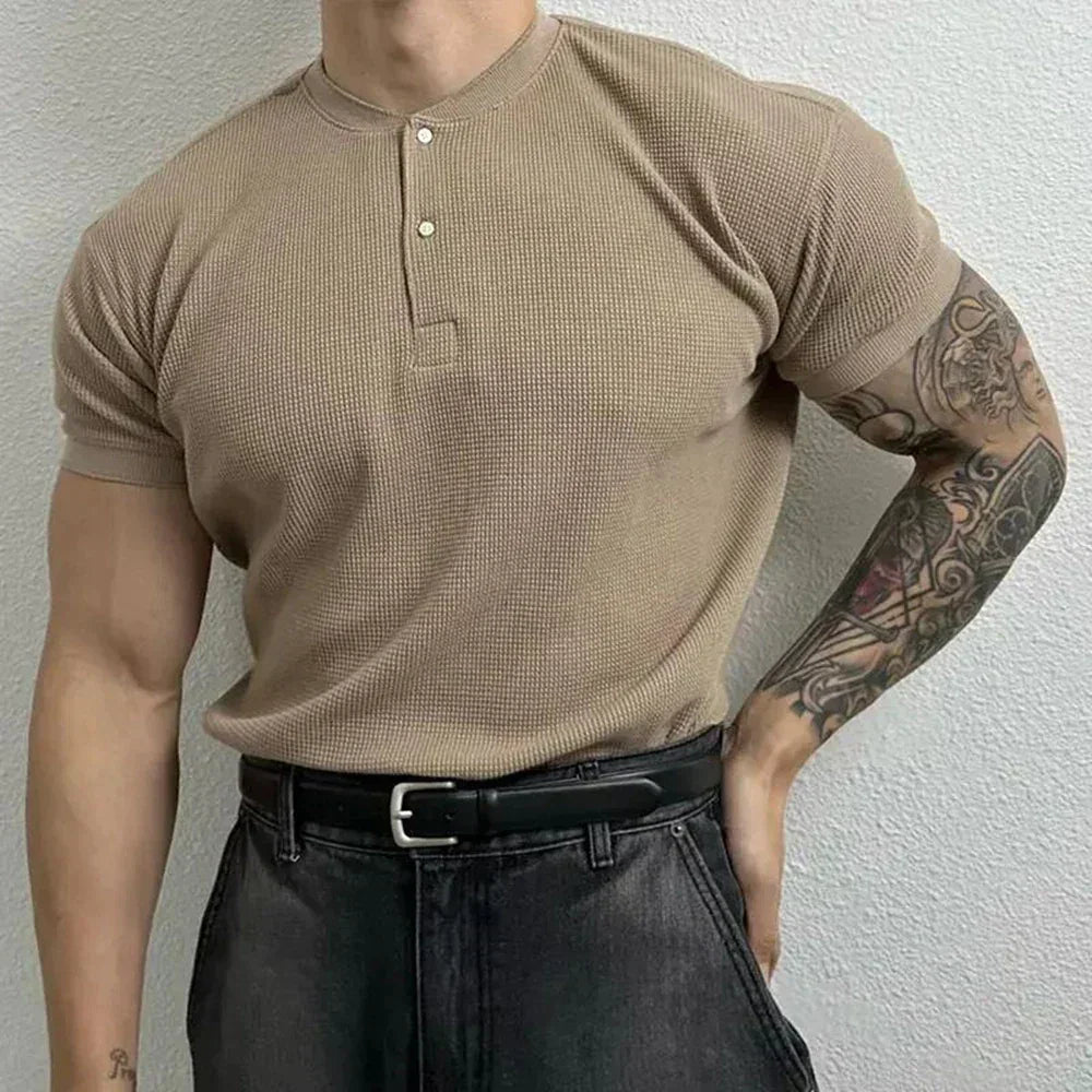 A muscular man wearing a form-fitting beige short-sleeve henley shirt with a Henley neckline, black belt, and dark jeans stands with one hand on his hip, showing a detailed sleeve tattoo on his right arm.