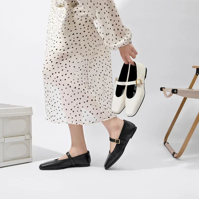 A person in a white dress with black polka dots wears black Mary Jane shoes and holds a pair of white Mary Jane shoes, standing beside a beige folding chair and a white chest, showcasing classic Mary Jane shoes style.