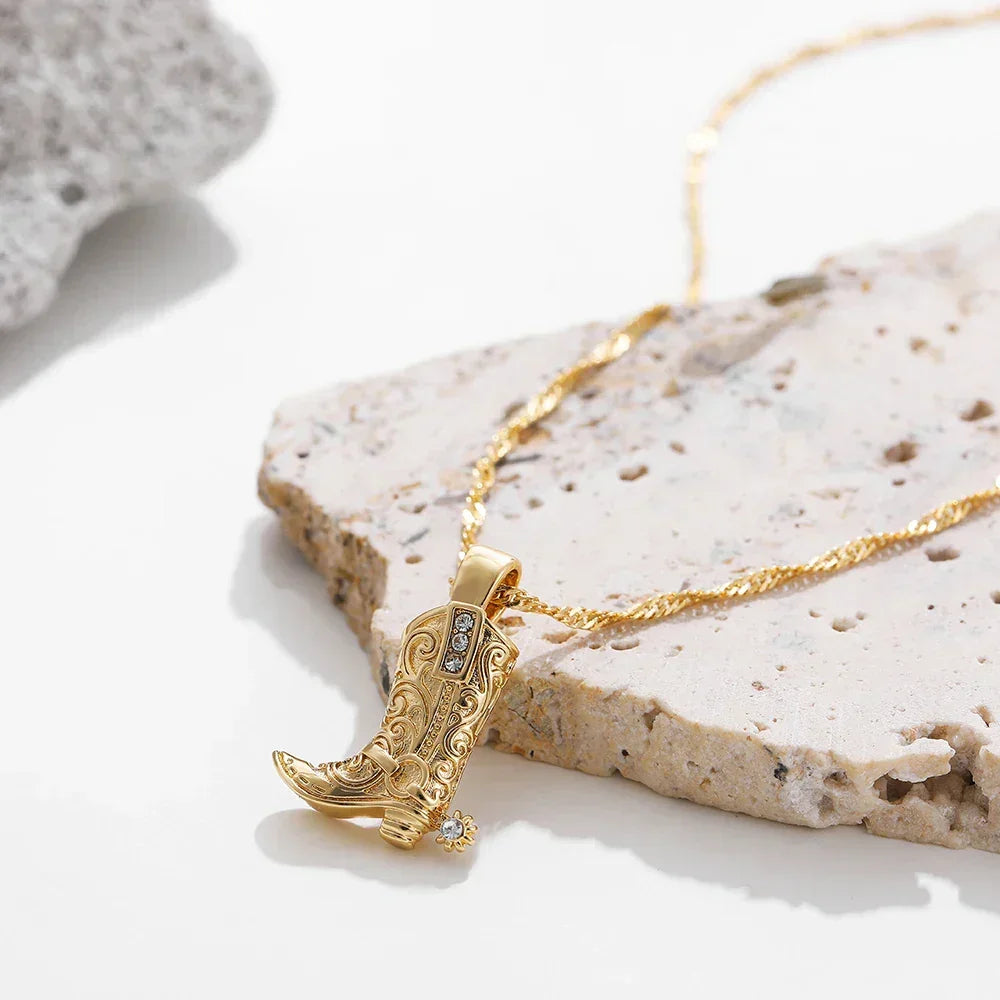 A gold cowgirl necklace with a detailed cowboy boot pendant, featuring decorative engravings and a small gemstone, displayed on a textured stone surface—a perfect piece of western jewelry.