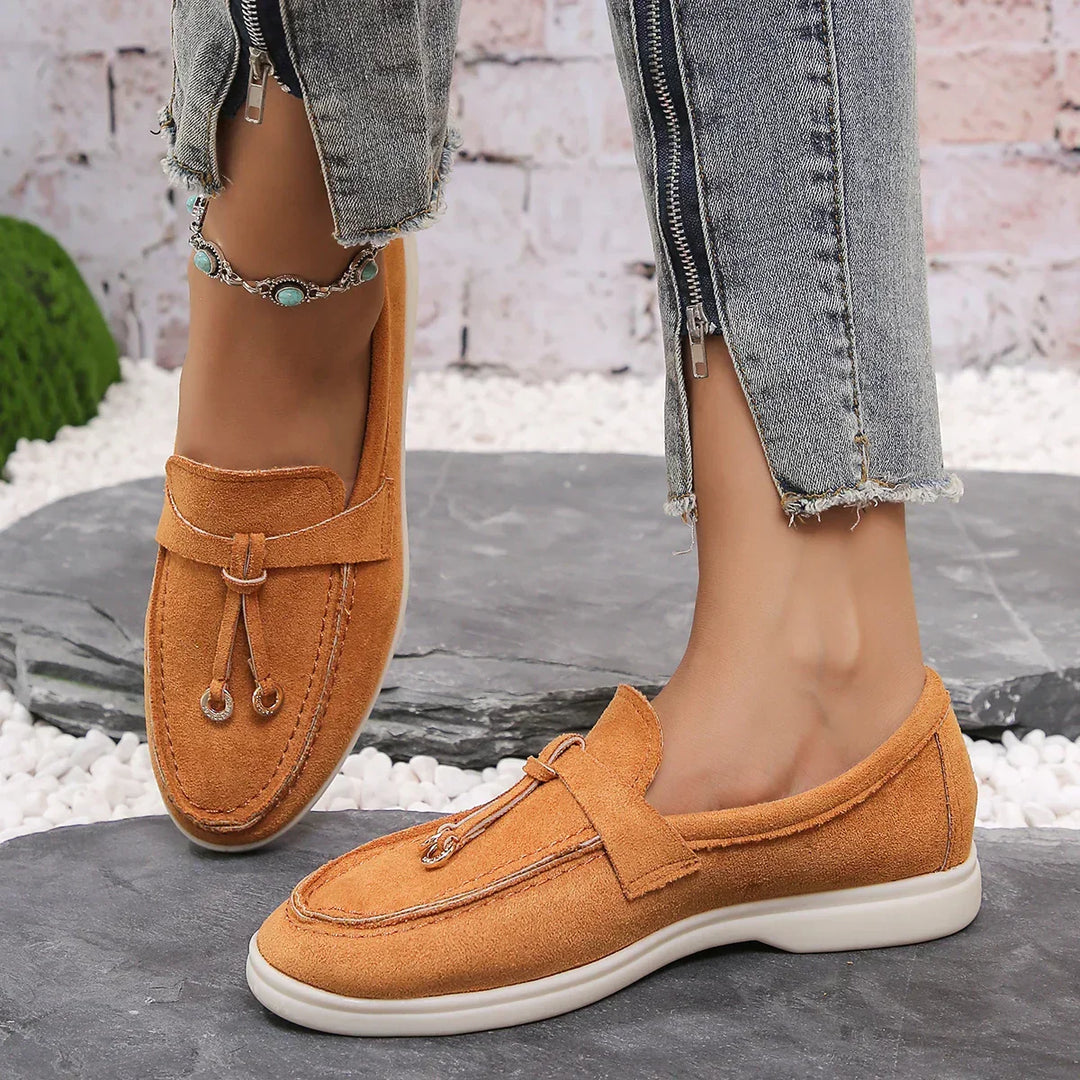 A person wearing light blue frayed-hem jeans and tan suede moccasins with white soles stands on a stone surface. One leg is crossed over the other, revealing an anklet with turquoise stones for a laid-back streetwear shoes vibe.