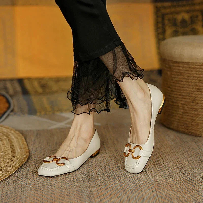 A person wearing cream-colored loafers with metallic accents and cushioned insole, paired with black pants with sheer ruffled hems, stands on a textured rug.