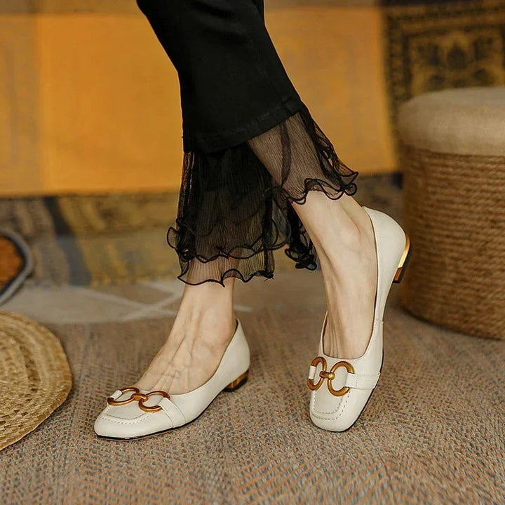 A person wearing cream-colored loafers with metallic accents and cushioned insole, paired with black pants with sheer ruffled hems, stands on a textured rug.
