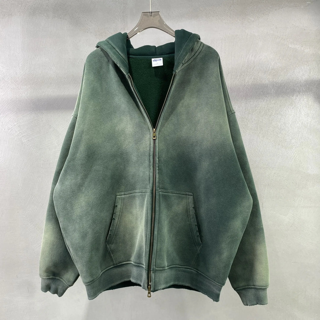 A dark green zip-up hoodie with a spray-dyed vintage, distressed look and relaxed fit hangs on a hanger against a light gray textured background.
