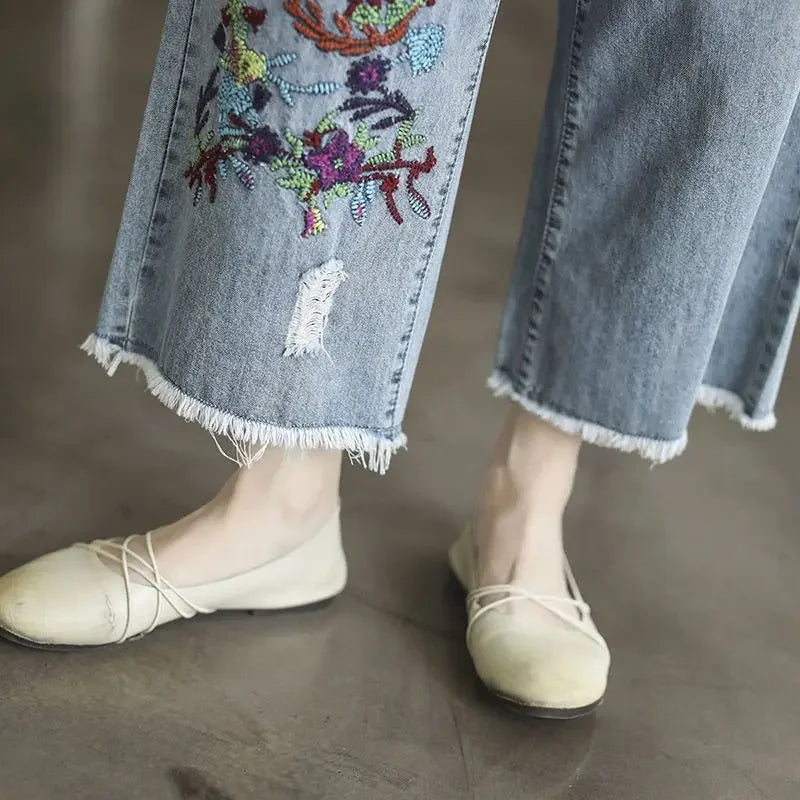 Person wearing light-colored flats and loose-fitting denim, wide-leg, frayed-hem jeans with colorful retro embroidery on one leg, standing on a smooth floor.
