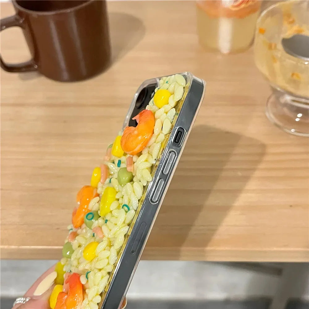 A hand holds a phone with a case decorated to look like colorful fried rice, featuring 3D detailing of imitation shrimp, peas, corn, and carrots. The shock-absorbent case rests on a wooden table next to mugs and dishes.