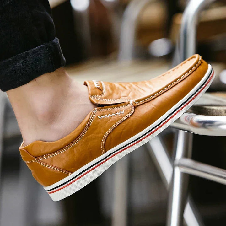 A person wearing light brown boat-style loafers with white stitching and a white rubber sole featuring red and black stripes, with their foot resting on a metal bar.