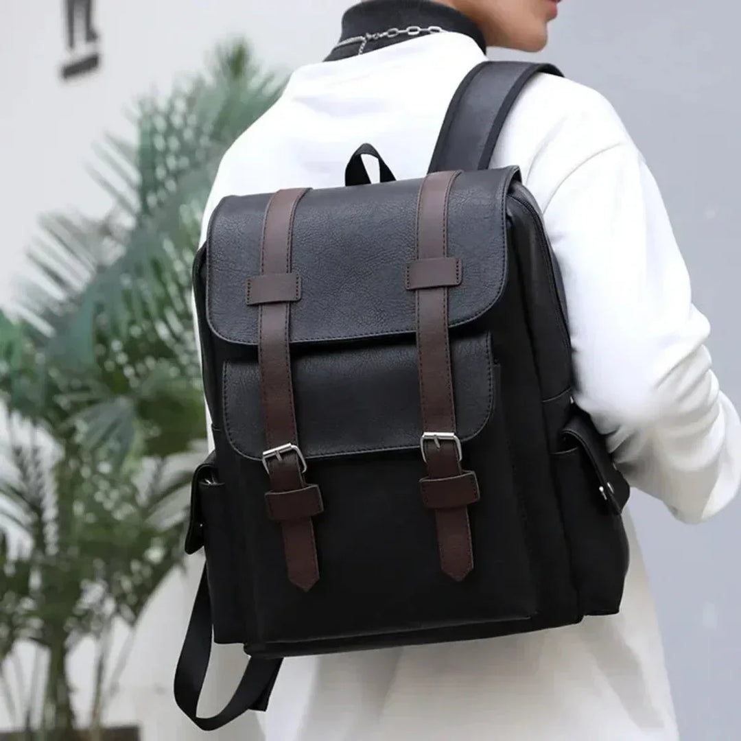 A person wearing a white jacket carries a black waterproof leather backpack with brown buckle straps on their back. The background features green plants and a light-colored wall.