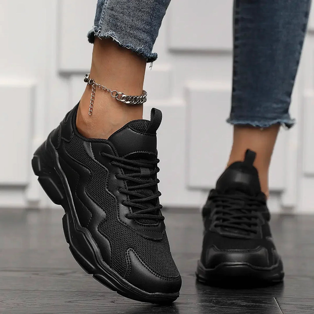 A person wearing black slip-on street shoes with advanced shock absorption and rolled-up jeans stands on a dark wooden floor, one foot slightly raised. The person is also wearing a silver chain anklet.