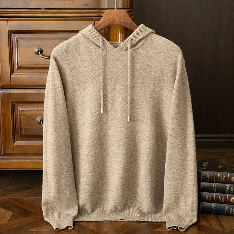 A beige cashmere blend hooded sweatshirt with drawstrings hangs on a wooden hanger, placed in front of a wooden dresser and next to a stack of books on a wooden floor. Perfect as a cozy layering sweater for chilly days.