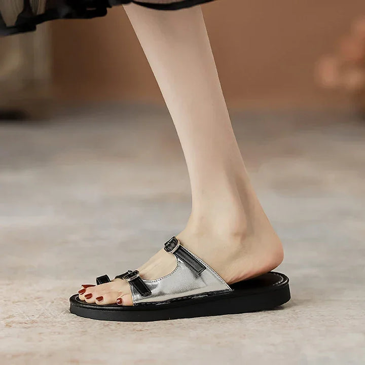 A person wearing a silver and black open-toe sandal with a leather upper and buckle strap, showing painted toenails, stands on a light-colored surface.