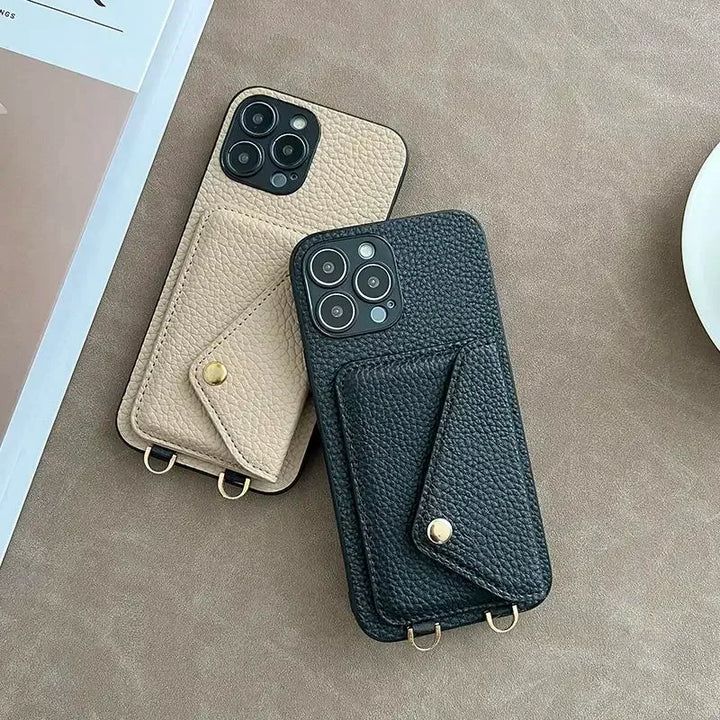 Two smartphones in beige and black pebble-texture material cases with attached cardholder pockets and gold button clasps rest on a soft brown surface beside a white saucer and a partially visible book or magazine.