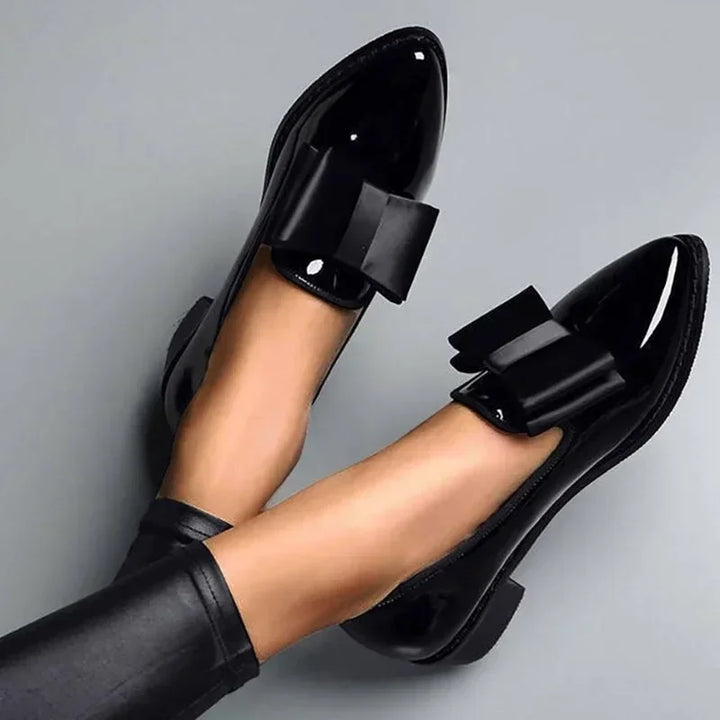 A person wearing shiny black patent leather loafers with statement bows and black leggings stands against a gray background.