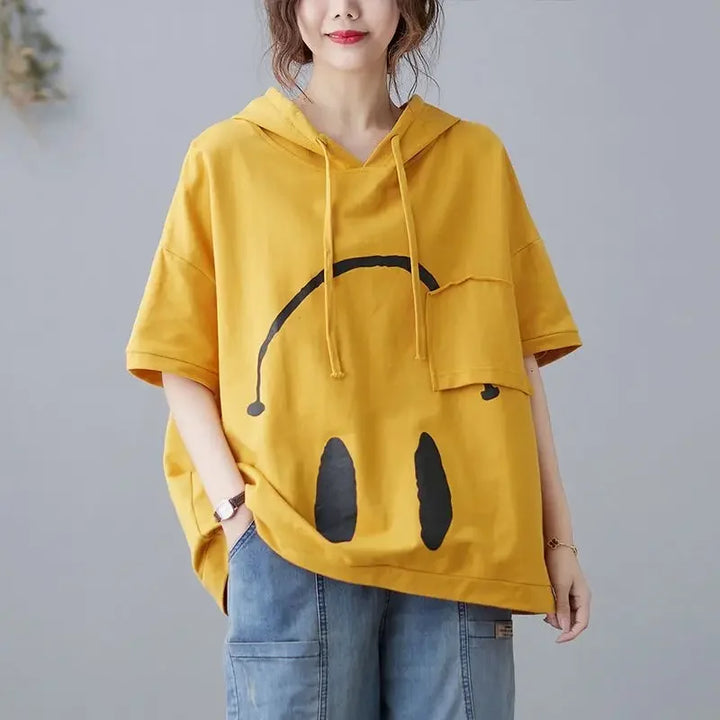 A person wearing a loose yellow Monday Mood hoodie with a large black abstract smiley face design and blue jeans, standing with hands in their pockets. The short-sleeve hoodie pops against the plain, light-colored background.