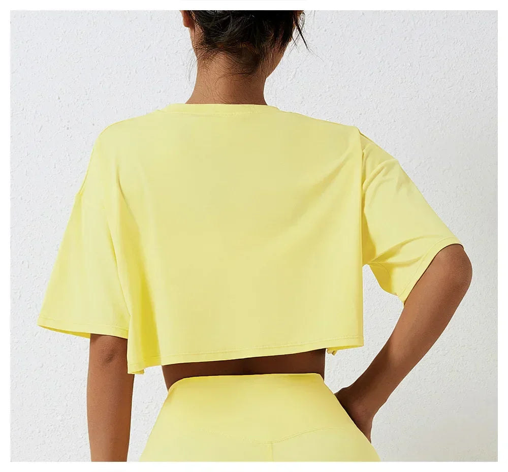 A person stands facing away from the camera, wearing a relaxed fit, short-sleeved yellow cotton crop top and matching high-rise pants, set against a white textured wall.