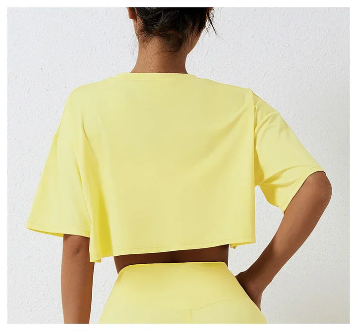 A person stands facing away from the camera, wearing a relaxed fit, short-sleeved yellow cotton crop top and matching high-rise pants, set against a white textured wall.