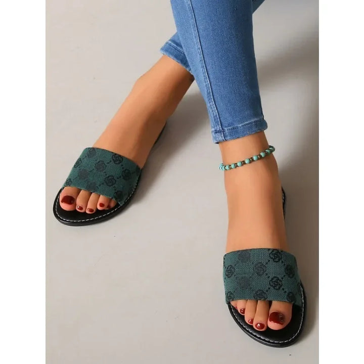 A person in blue skinny jeans and green open-toe modern slippers with a subtle pattern, plus a turquoise beaded anklet, sits with legs crossed on a light-colored floor—a perfect touch of summer and British style.